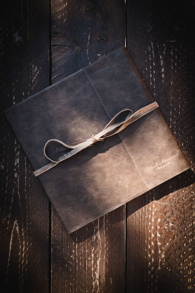 Hand-stitched natural Tuscany leather wedding photo album tied with ribbon, resting on dark wood — Thibault Maestracci Photographer