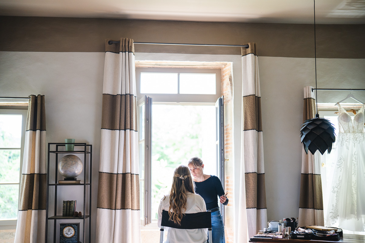 The bride by a window in contre-jour light during preparations at La Bélonie, Tarn