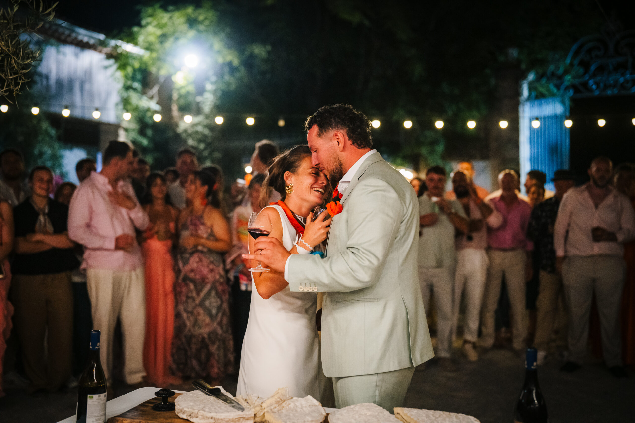 The bride and groom dancing close together in front of their guests under string lights at an evening wedding party