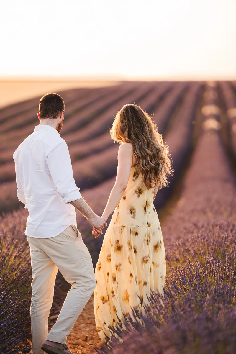 A newly engaged couple walking away hand in hand through a lavender field