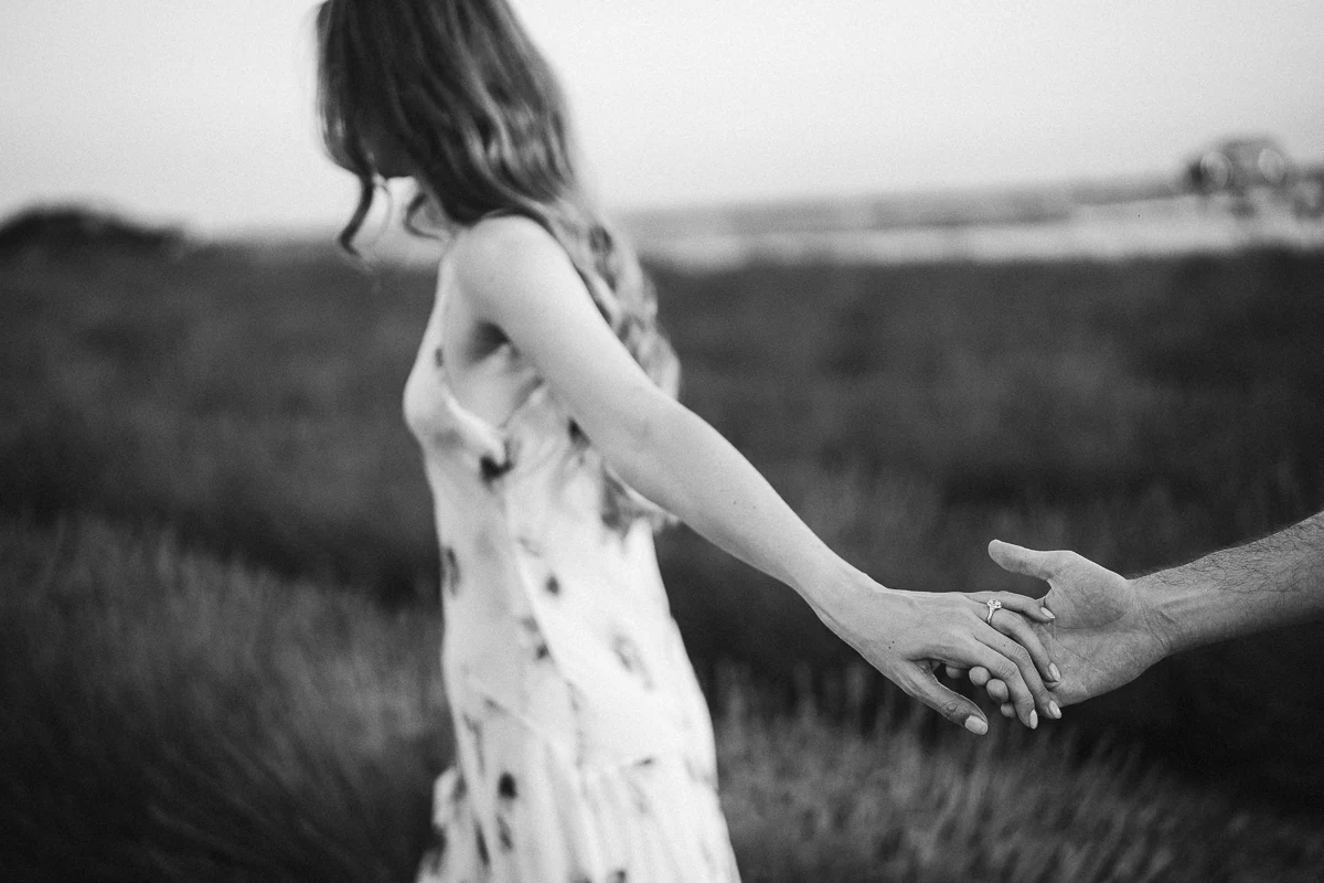 A woman leading her partner by the hand through a lavender field, engagement ring visible, black and white