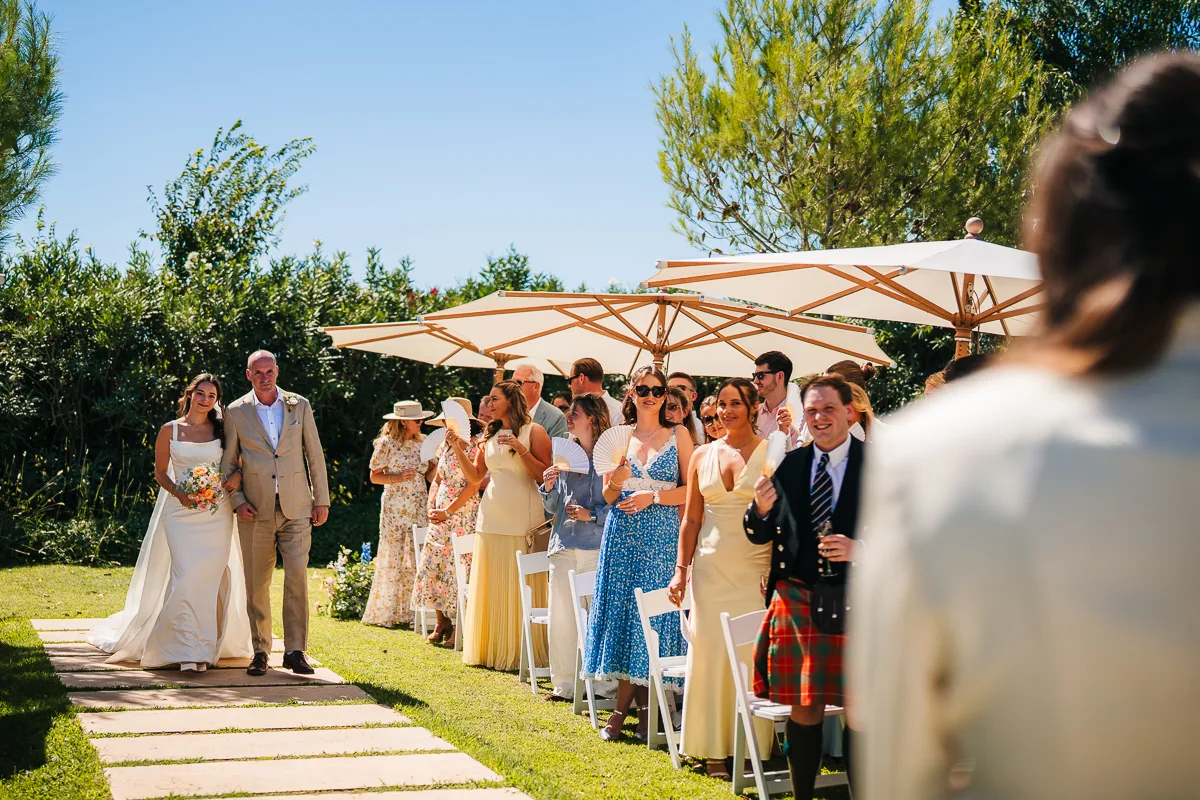 Bride walking down the garden aisle with her father between guests under parasols, Domaine de la Trinité