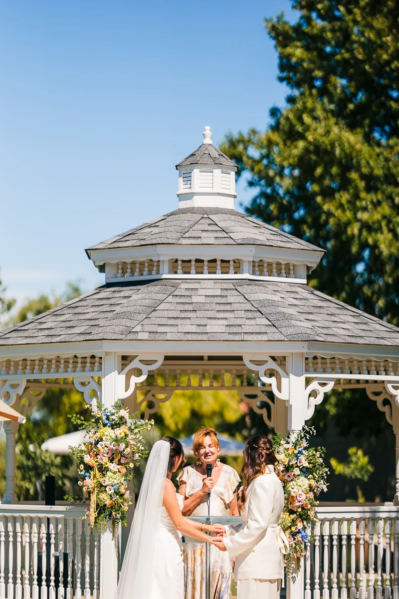 Two brides holding hands in front of the officiant in the white bandstand at Domaine de la Trinité