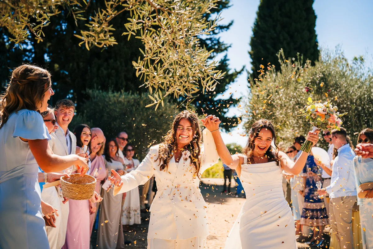 Two brides walking out of their ceremony through confetti and cheering guests under olive trees