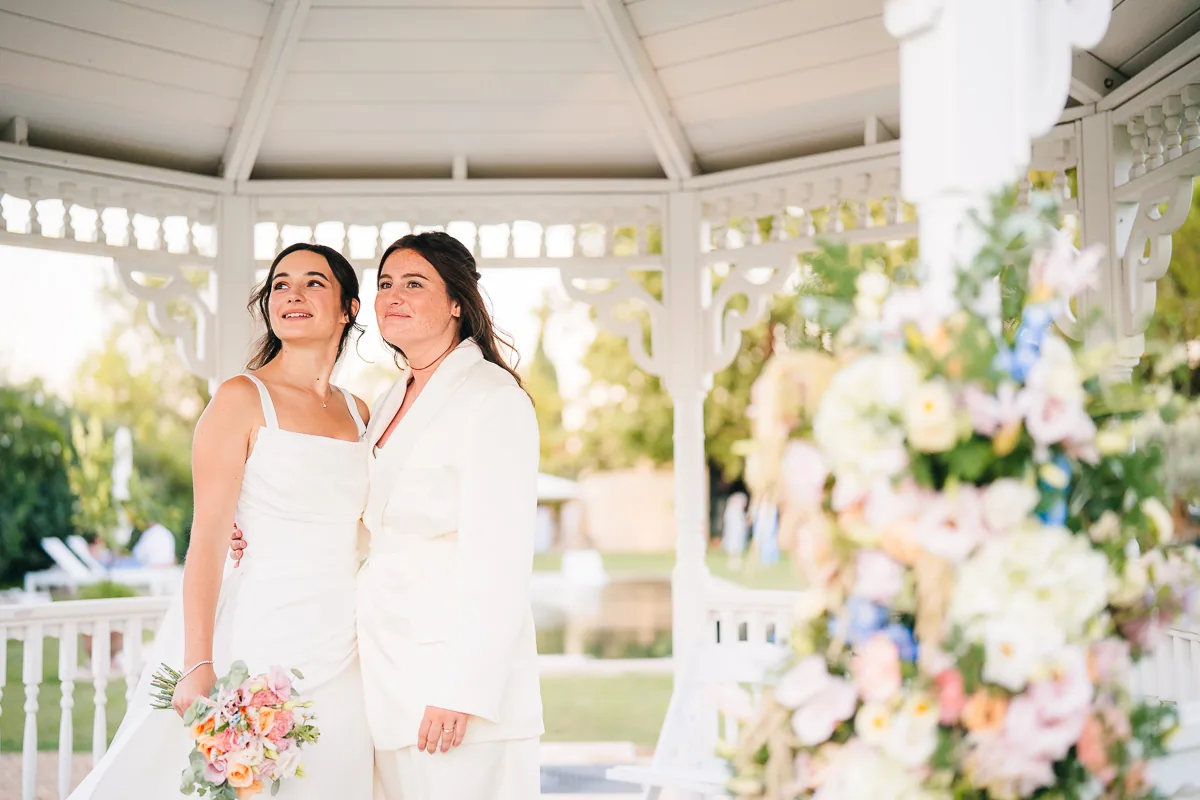 Two brides embracing in the white bandstand with pastel flowers at Domaine de la Trinité