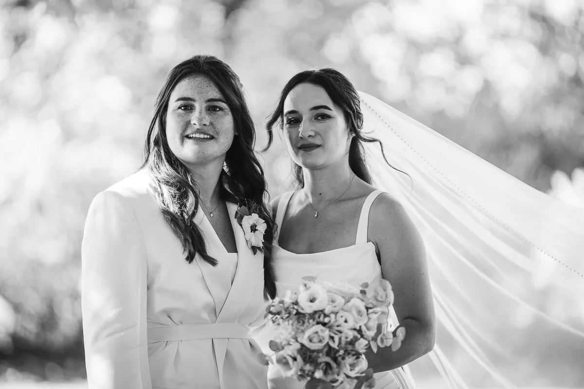 Black and white portrait of two brides, one in a white suit and one in a dress with a long veil
