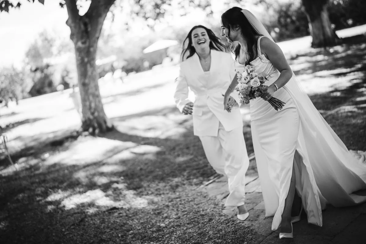 Black and white photo of two brides running and laughing under the trees on their wedding day