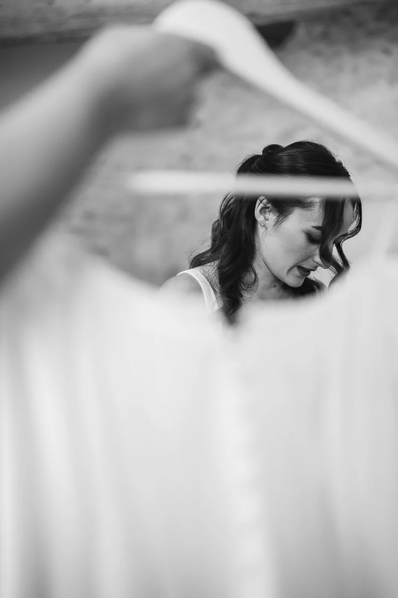 Black and white portrait of a bride framed by the wedding dress and its hanger while getting ready