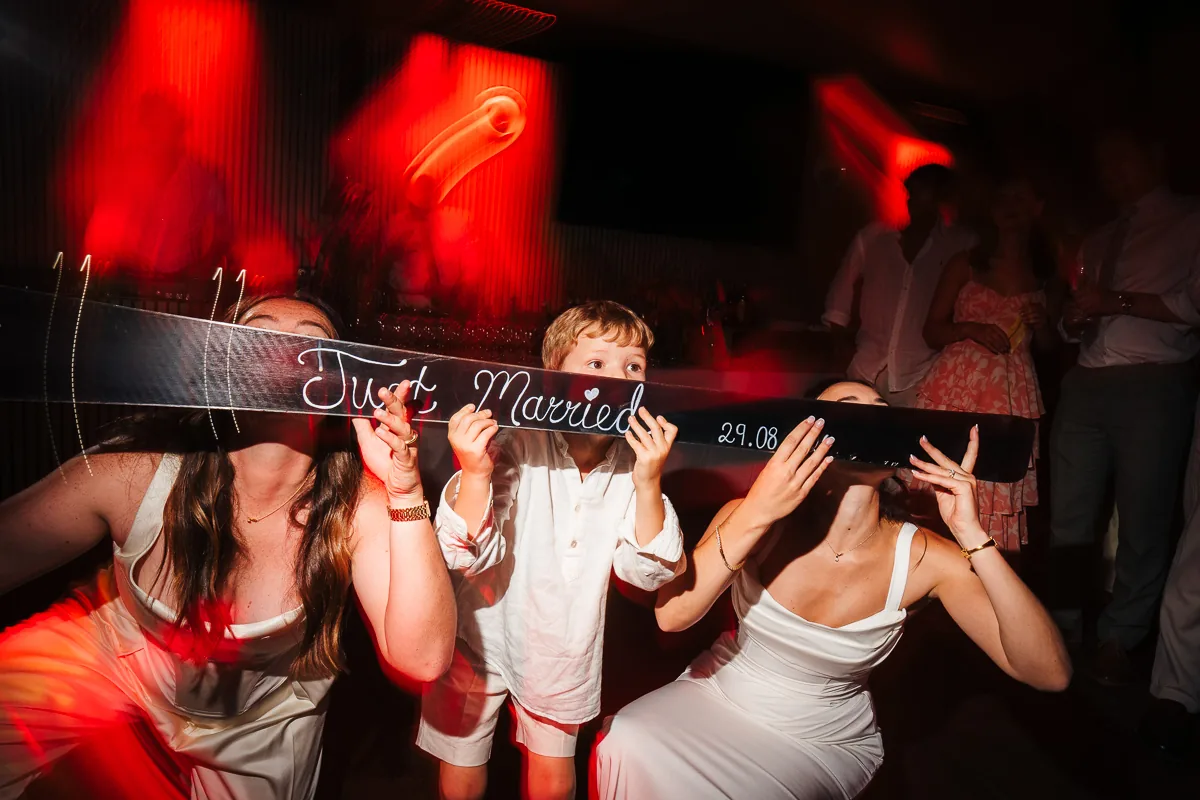 Two brides and a young guest holding a Just Married sign on the dance floor, Domaine de la Trinité