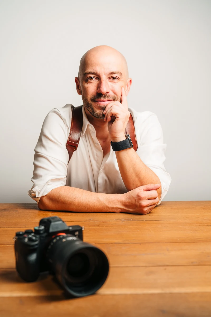 Thibault Maestracci, wedding photographer, chin on hand behind his camera on a wooden table
