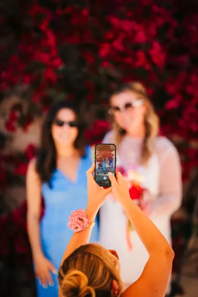 Guest photographing the bride and a fellow guest on a phone, with blurred flowers and colorful light in the foreground