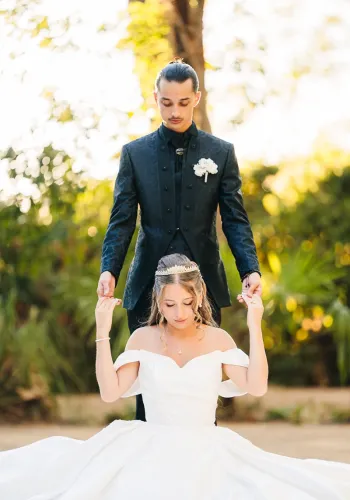 A groom in a dark patterned suit stands behind his seated bride in a white gown, holding her raised hands, in the garden of Domaine de l'Argentière near Montblanc